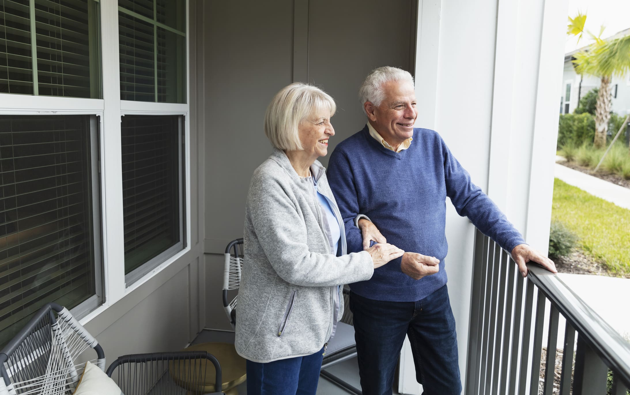 A senior couple in their 70s standing together on their apartment balcony. They are looking at the view, smiling. The woman is holding her husband's arm.